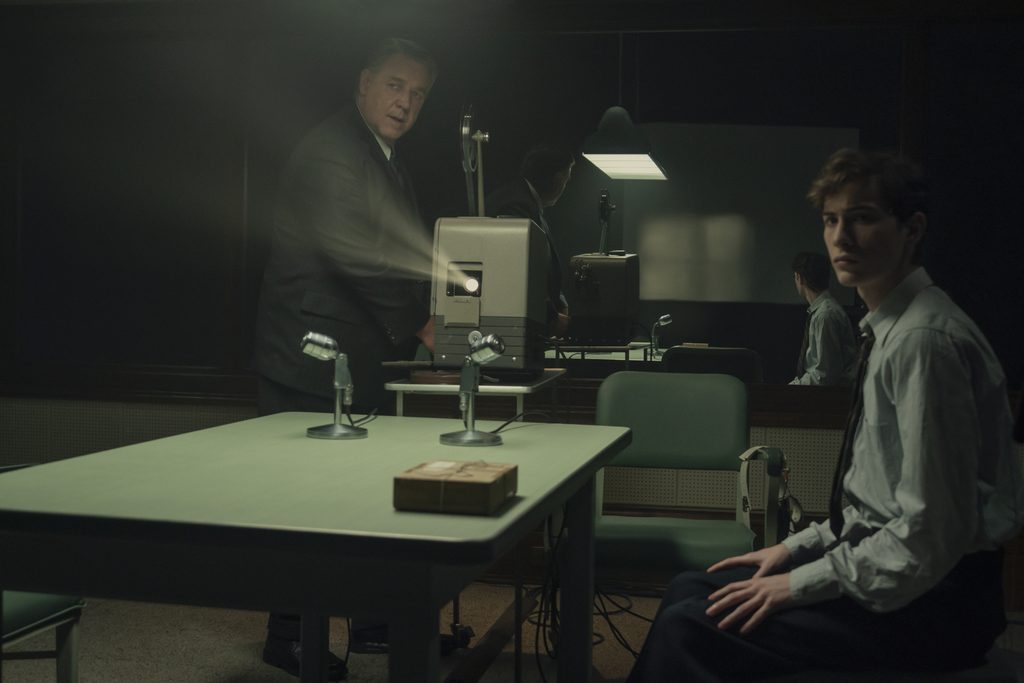"Two men in formal attire are in a dimly lit interrogation room with a green table, microphones, and a film projector. One man stands by the projector while the other sits, suggesting a tense interview or investigation setting."